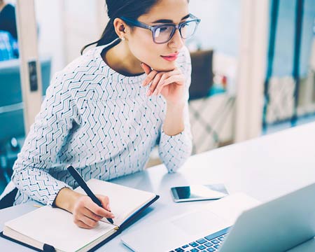 Woman in a pony tail wearing glasses looking at a laptop and taking notes