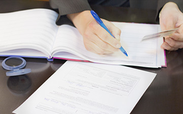 Photo of an open Notary hournal with papers around it on a desk. A person's hands are shown holding a pen in one hand and an ID in the other.