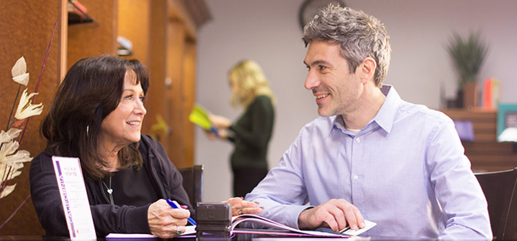 Photo of a man and woman, seated, looking at each other. There is an open notary journal and a stamp sitting on the desk in front of them. A blurred image of a woman reading a book is behind them.