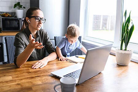 A woman working from home with a laptop, as a child sits next to her drawing at the table.