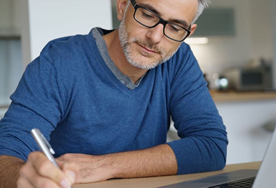 Man wearing glasses writing in a notebook while working on a laptop.