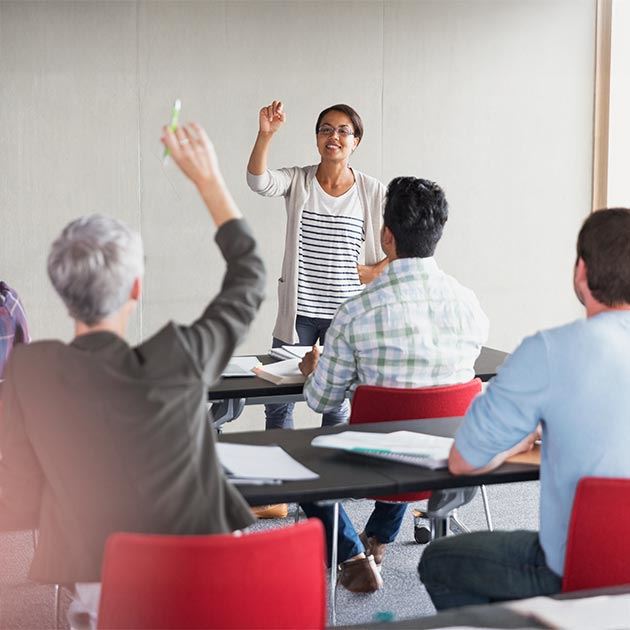 Students with their backs in view looking at a female instructor holding her hand up to call on a woman seated at a desk