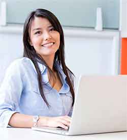 Woman with long dark hair in front of a laptop looking at the camera and smiling